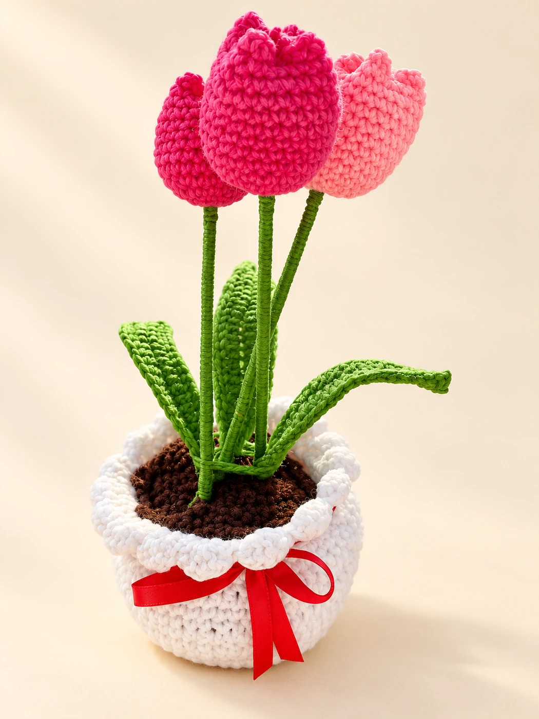 Pink crochet tulips with green leaves in a white pot with a red bow on a cream background