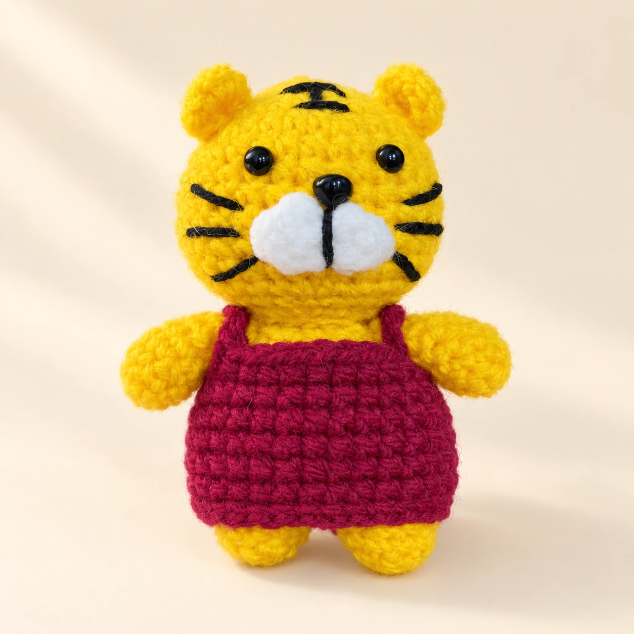 A yellow tiger crochet plush with black stripes and a burgundy dress on a cream background
