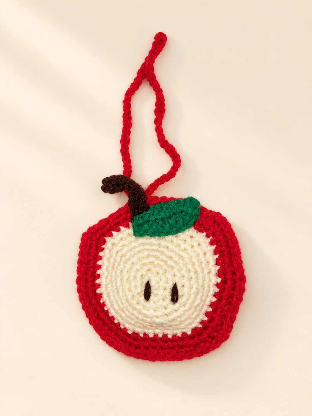 A red apple-shaped crochet case with a cream center, green leaf and red hanging loop on a cream background