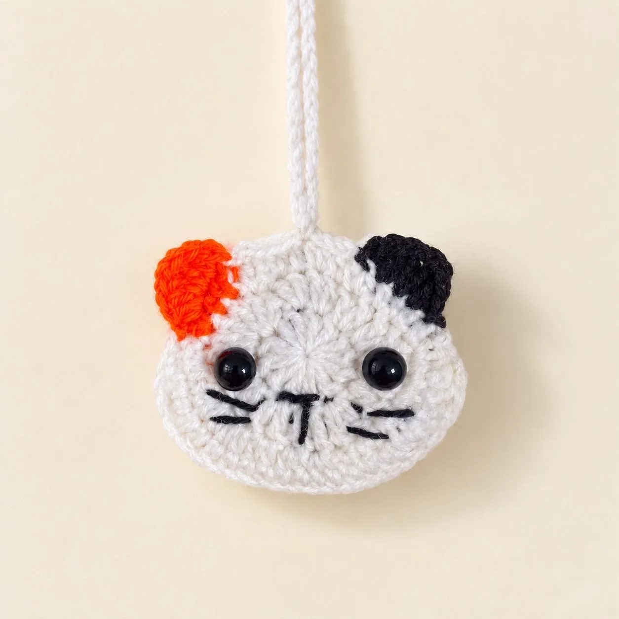 A white crochet cat-face earbud pouch with one orange and one black ear and a white strap