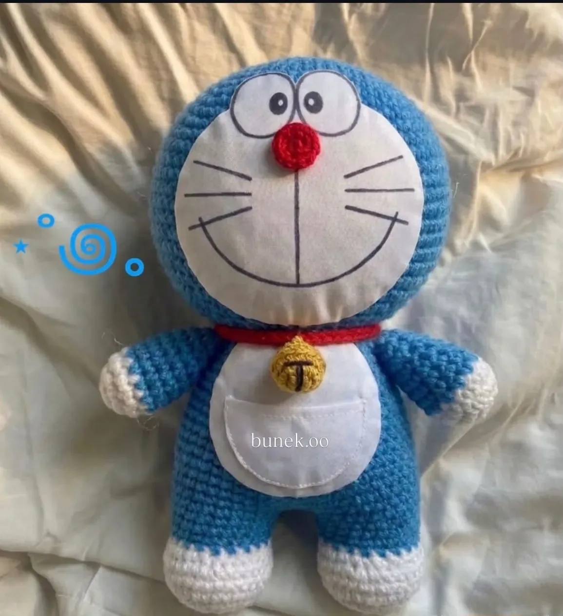 A blue and white crochet Doraemon plush with a red collar, golden bell and white pocket, lying on a cream bedsheet