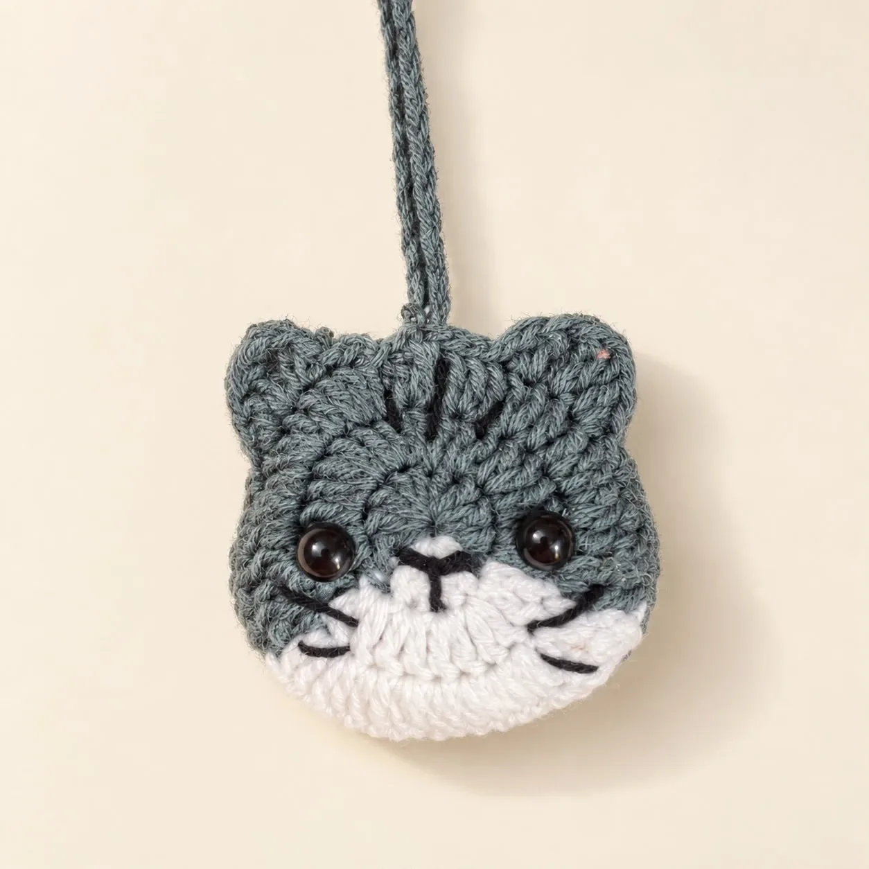 A grey crochet cat-face earbud pouch with black eyes, a white muzzle and a long grey strap
