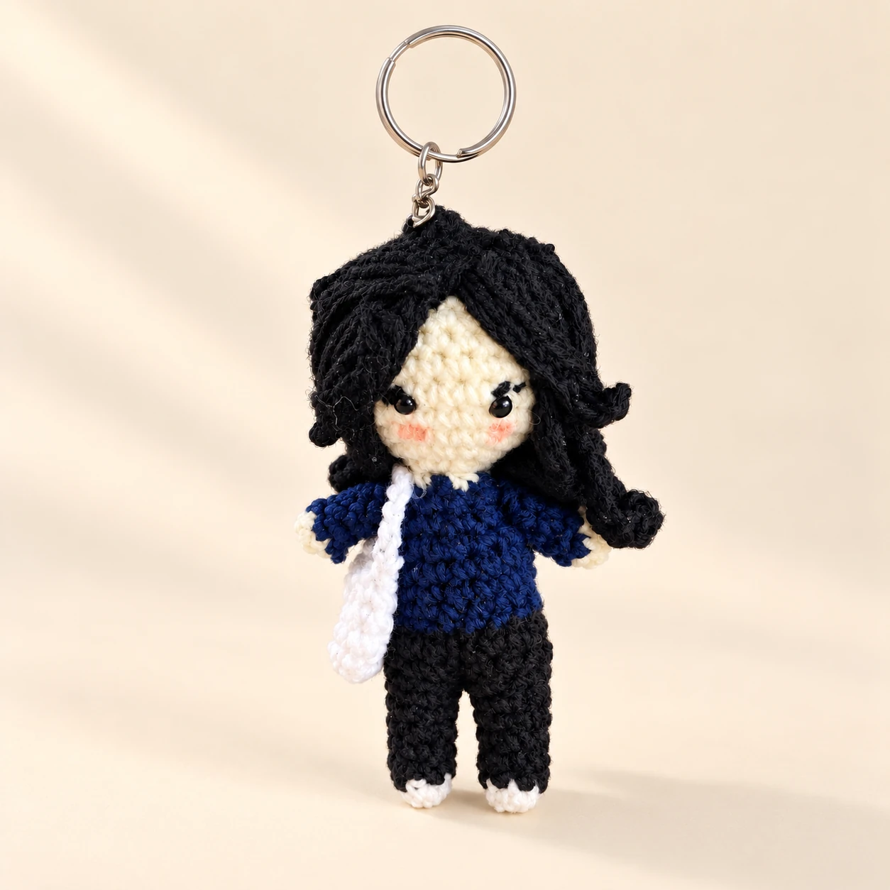 A black-haired crochet doll keychain in a navy top and black trousers with a white shoulder bag on a cream background