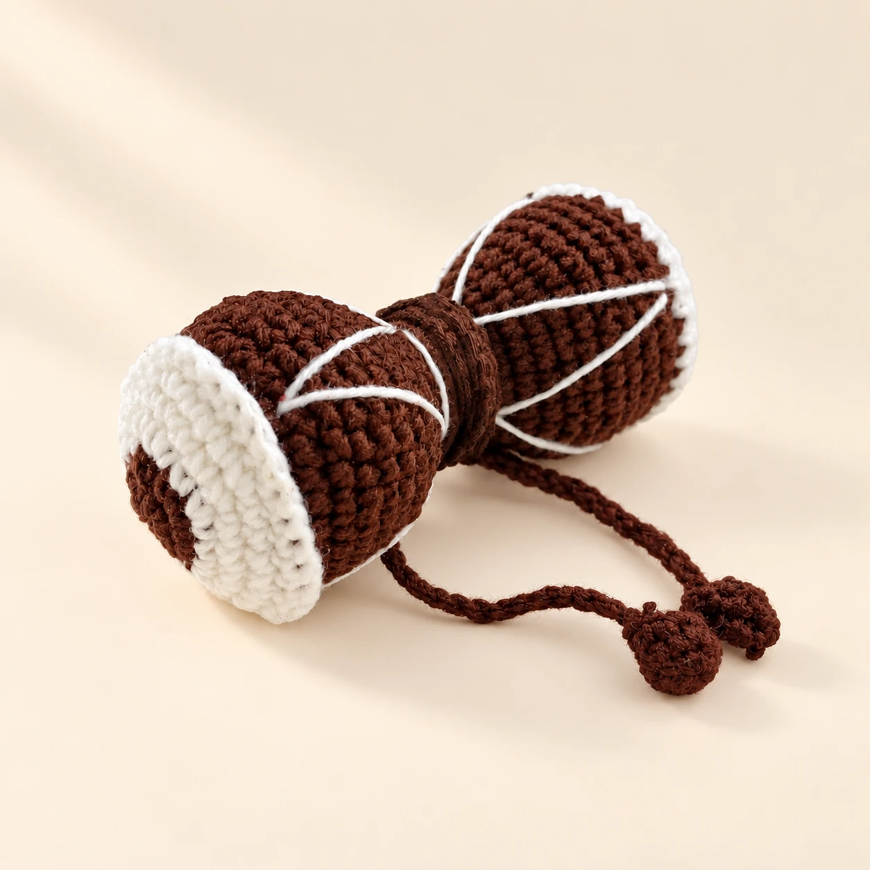 A brown crochet damaru with white rims, crossed white strings and hanging cords on a cream background