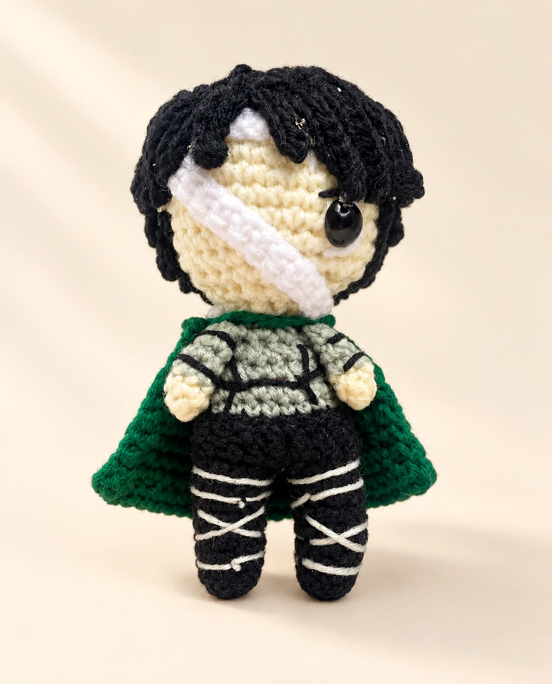 A Levi Ackerman crochet doll with black hair, a white face bandage and green cloak on a cream background