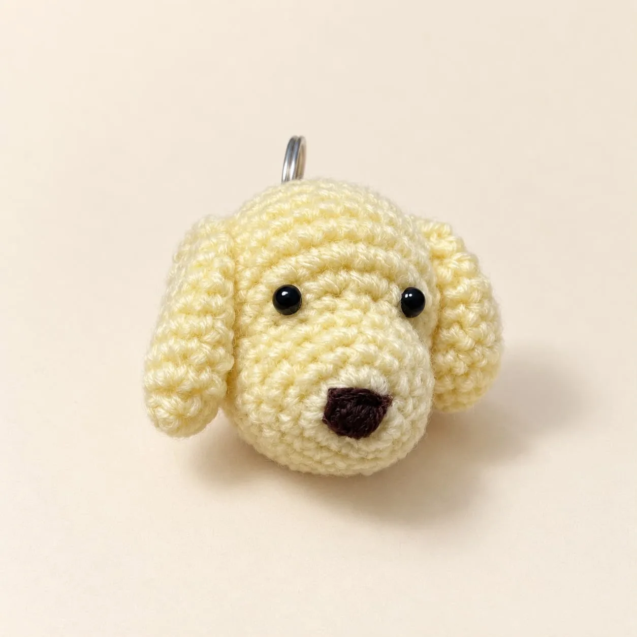 A pale yellow crochet puppy head keychain with floppy ears, black eyes and a brown nose