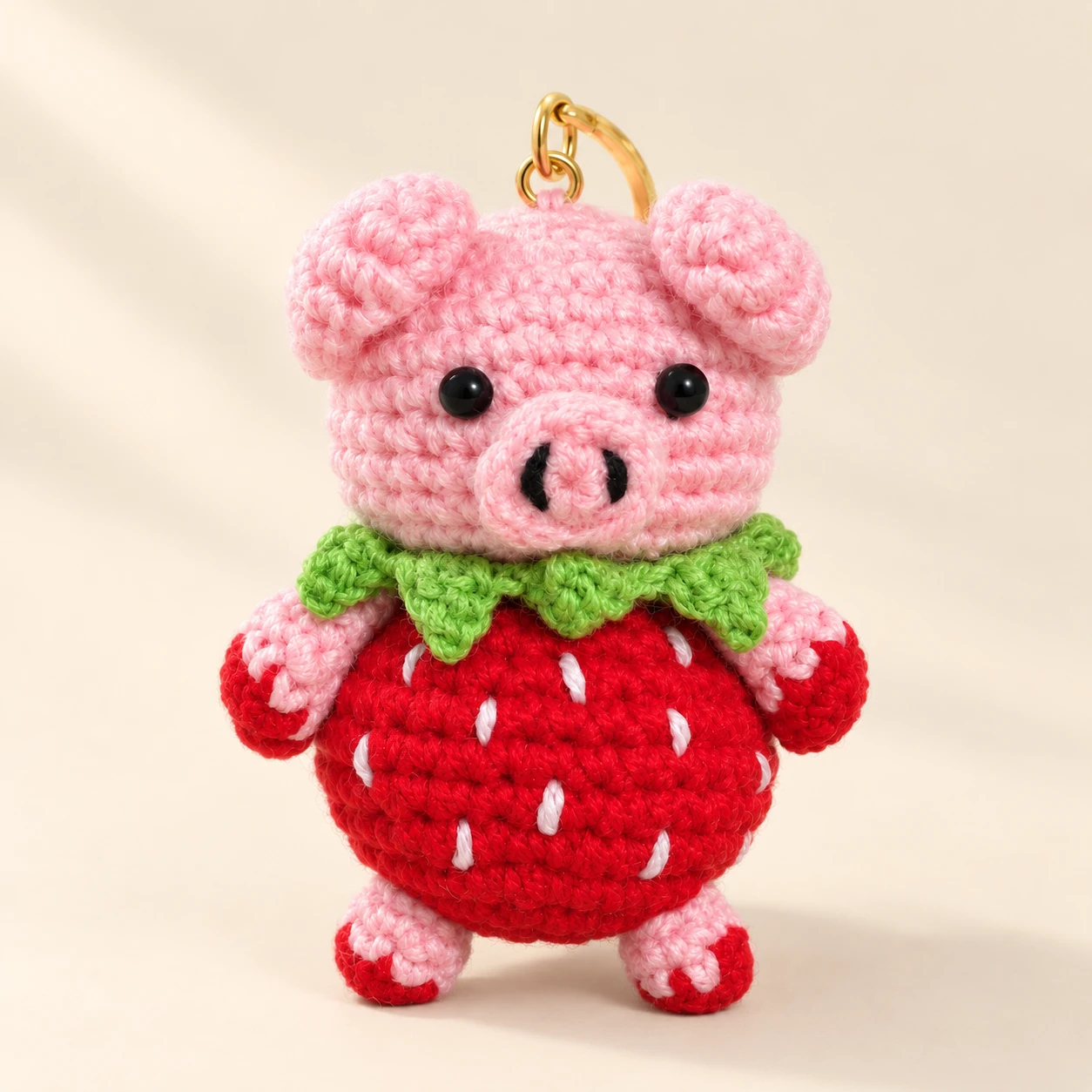 A pink crochet pig keychain in a red strawberry outfit with green leaves on a cream background