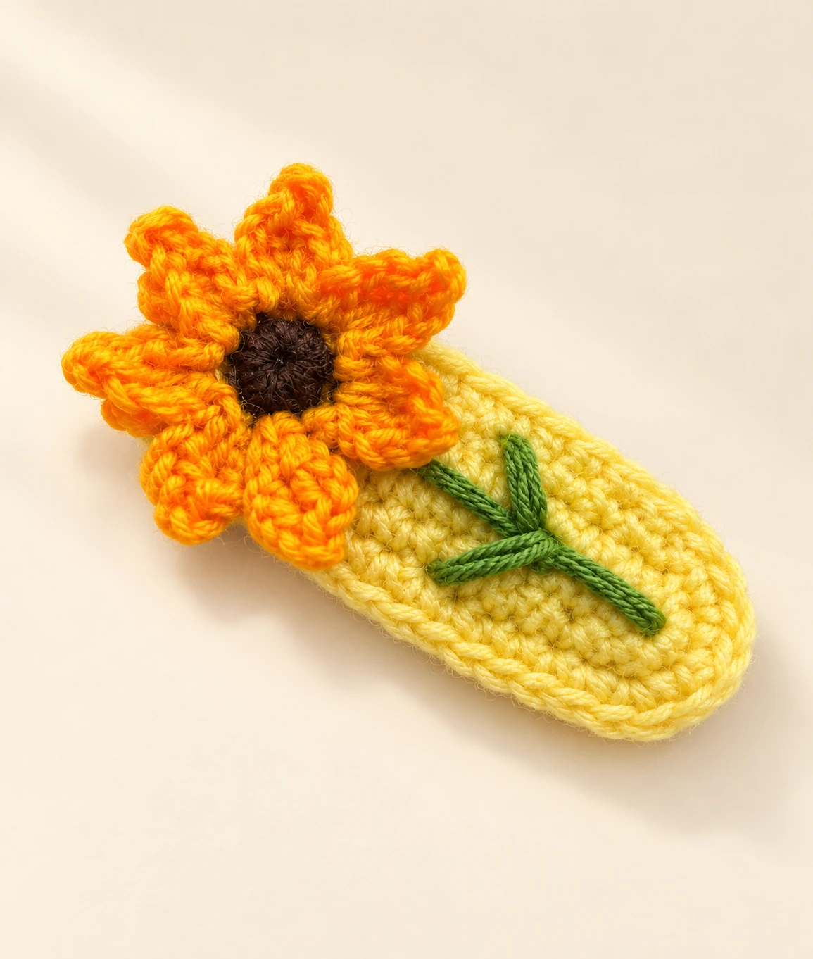 A yellow crochet sunflower hair clip with a green stem and leaf on a cream background