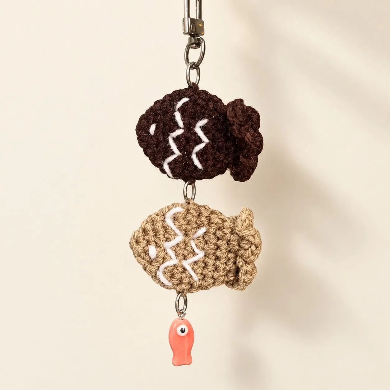 A keychain of two crochet taiyaki fish, one dark brown and one tan with white stitching, above a small coral fish charm