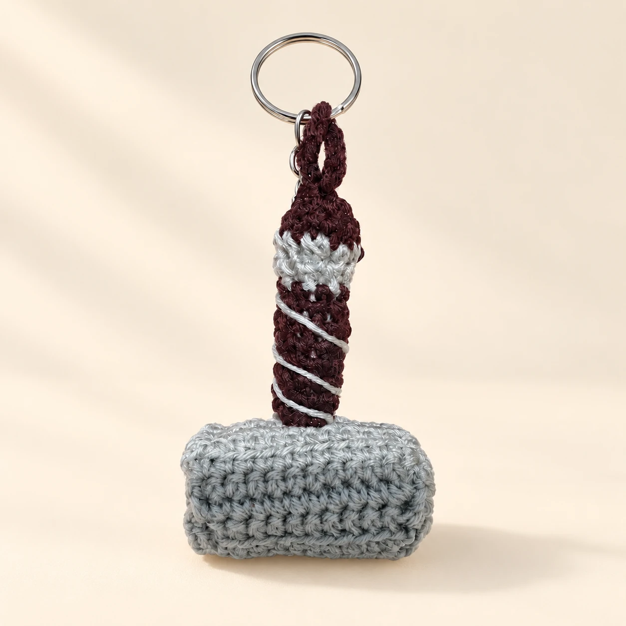 A gray crochet hammer keychain with a brown wrapped handle and metal ring on a cream background